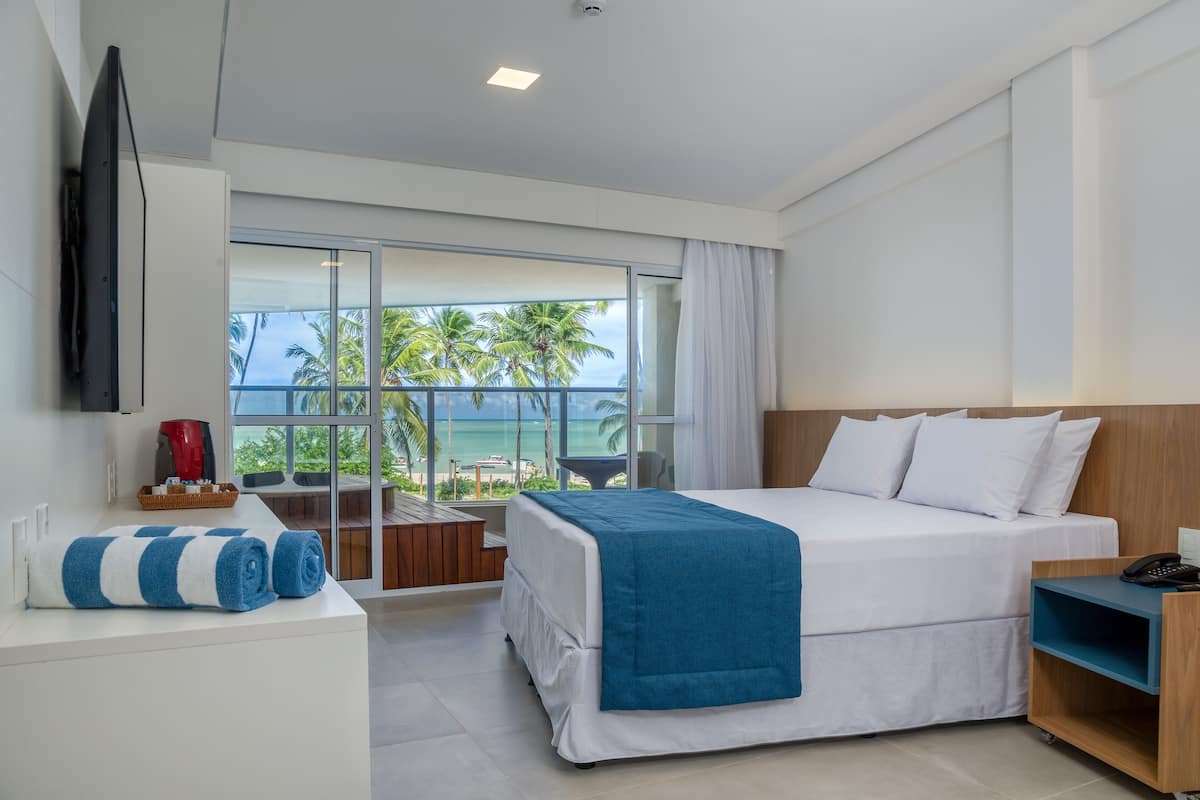 Een moderne hotelkamer met een groot bed, een aan de muur bevestigde televisie, een balkon met uitzicht op palmbomen en de oceaan, en een bureau met een koffiezetapparaat.