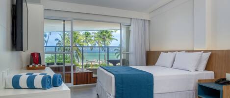 A modern hotel room with a large bed, a TV mounted on the wall, a balcony with a view of palm trees and the ocean, and a desk with a coffee maker.