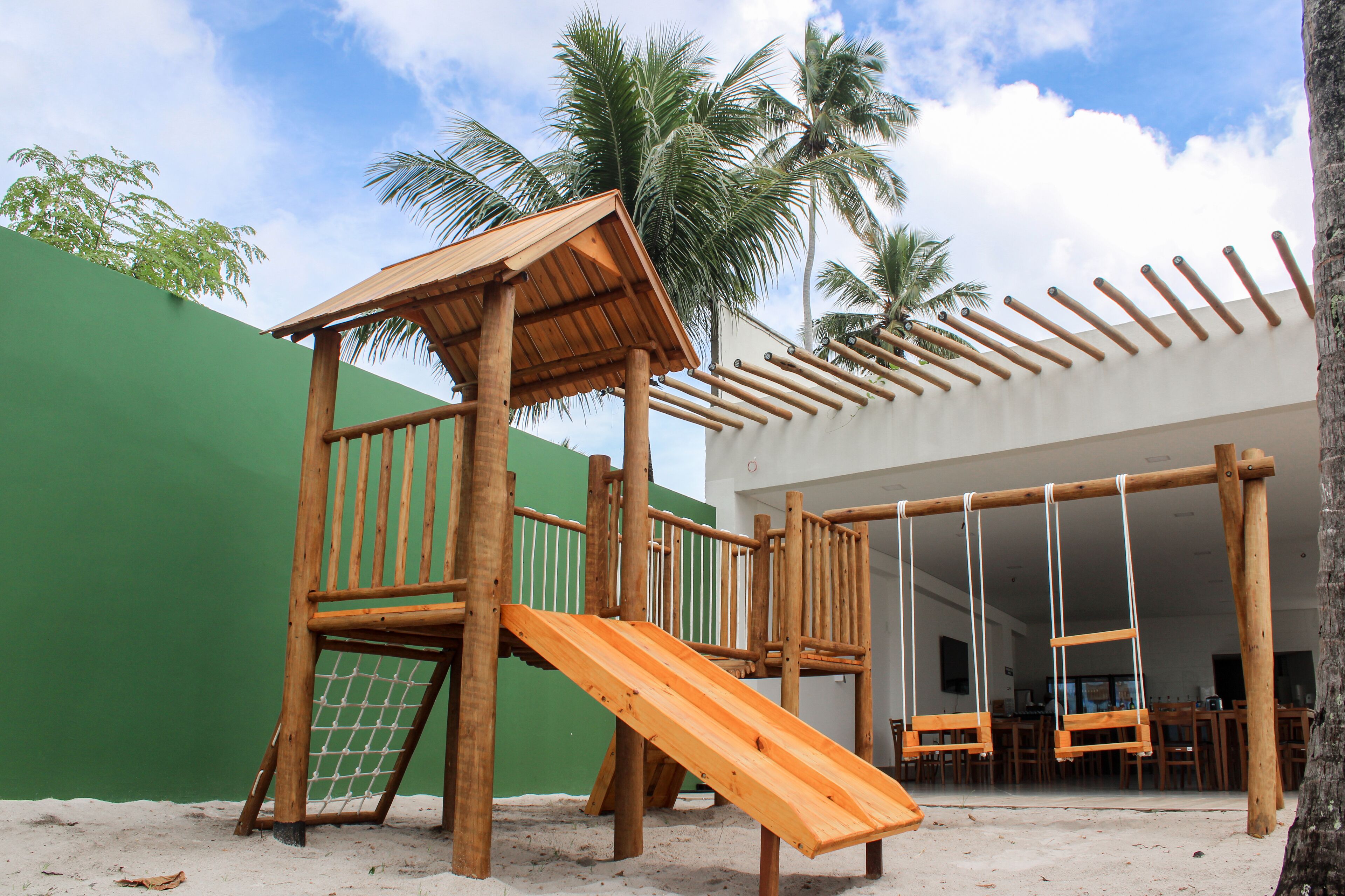 A wooden play structure with a slide and swings, set against a green wall and palm trees.
