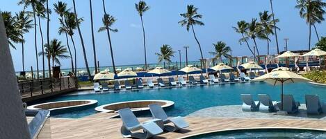 A resort pool area with lounge chairs and umbrellas, surrounded by palm trees and a clear sky.
