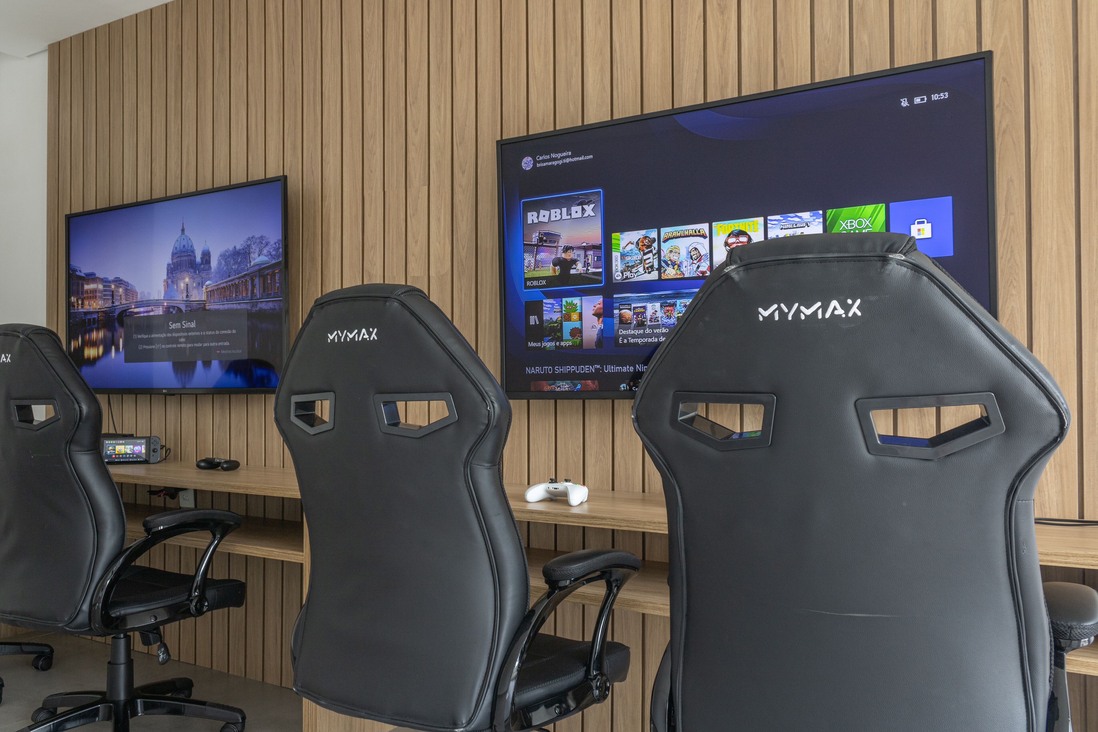 A gaming setup with three Mymax chairs, a large TV displaying a cityscape and a Roblox game, and a wooden wall.