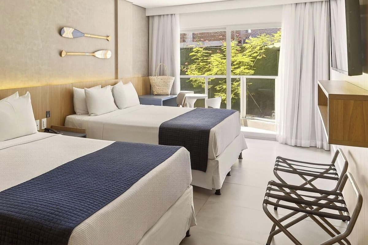 Een hotelkamer met twee bedden, een bureau, een stoel en uitzicht op groen.