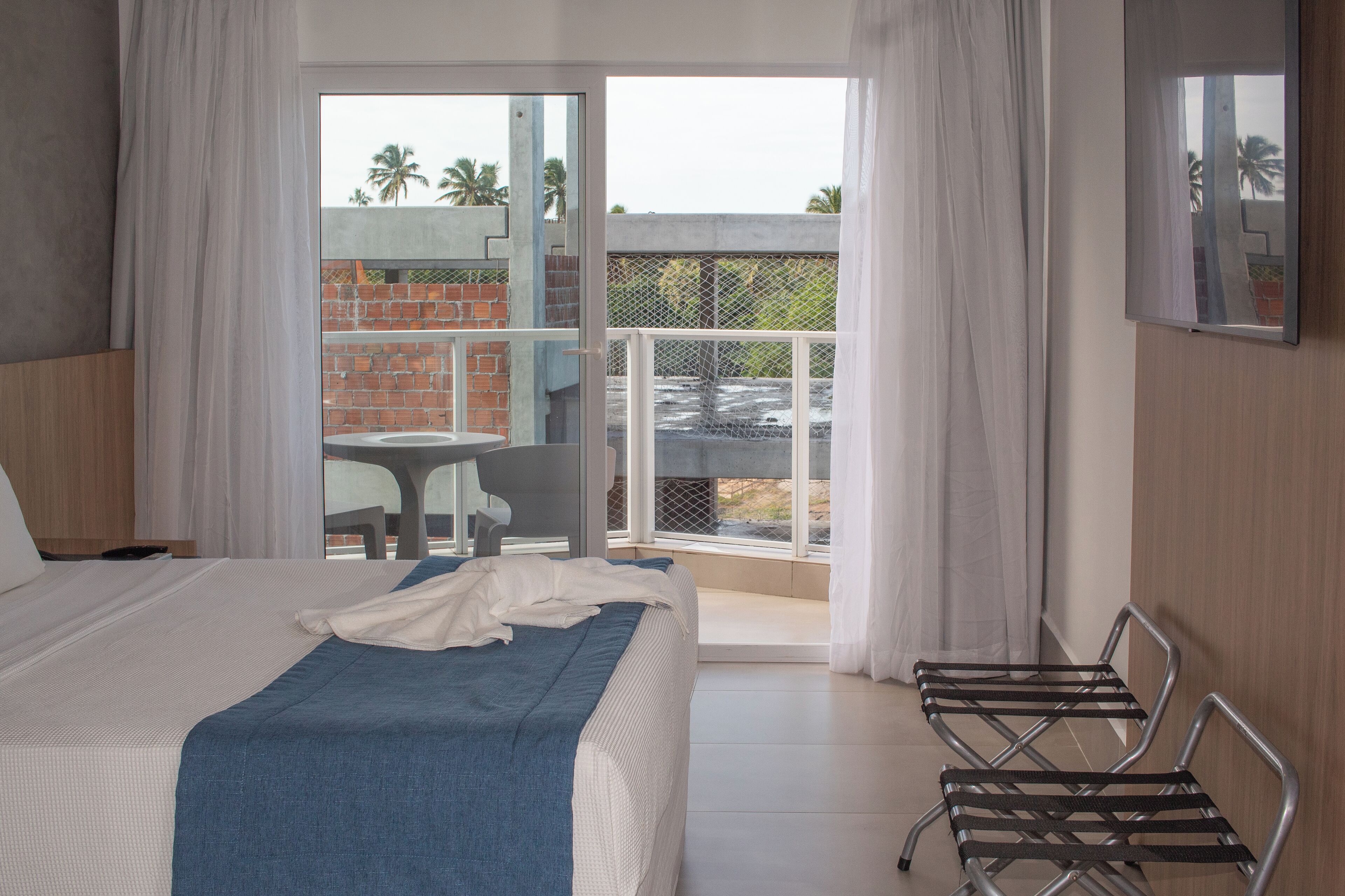 A hotel room with a bed, a chair, a balcony with a table and chairs, and a view of palm trees and a brick building.
