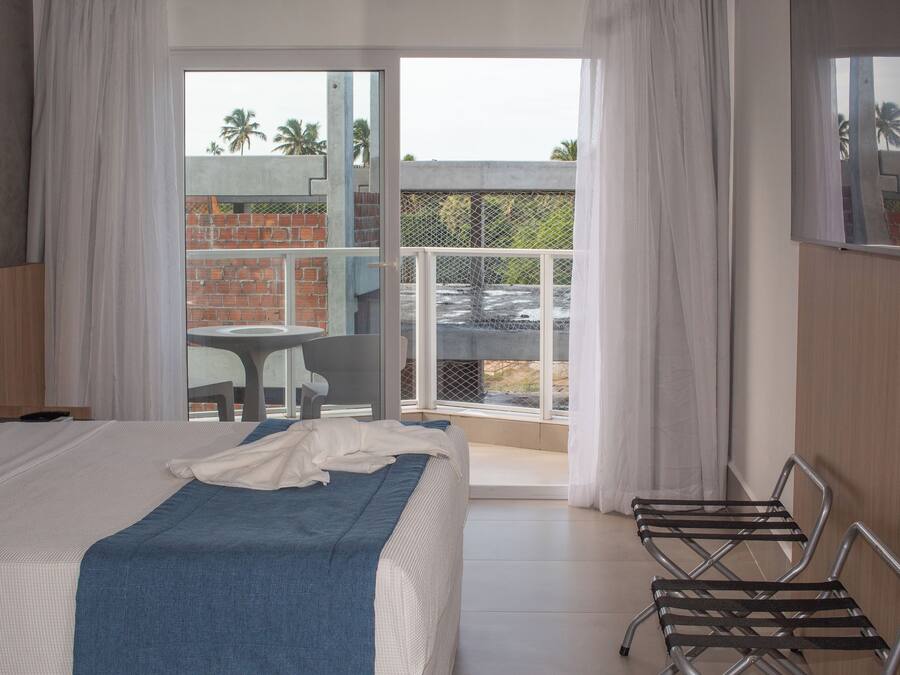 A hotel room with a bed, a chair, a balcony with a table and chairs, and a view of palm trees and a brick building.
