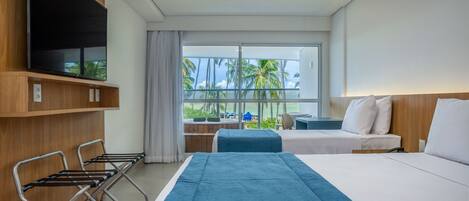 Habitación de hotel con una cama grande, un televisor montado en la pared, un balcón con vistas a palmeras y una zona de estar con una mesa y sillas.