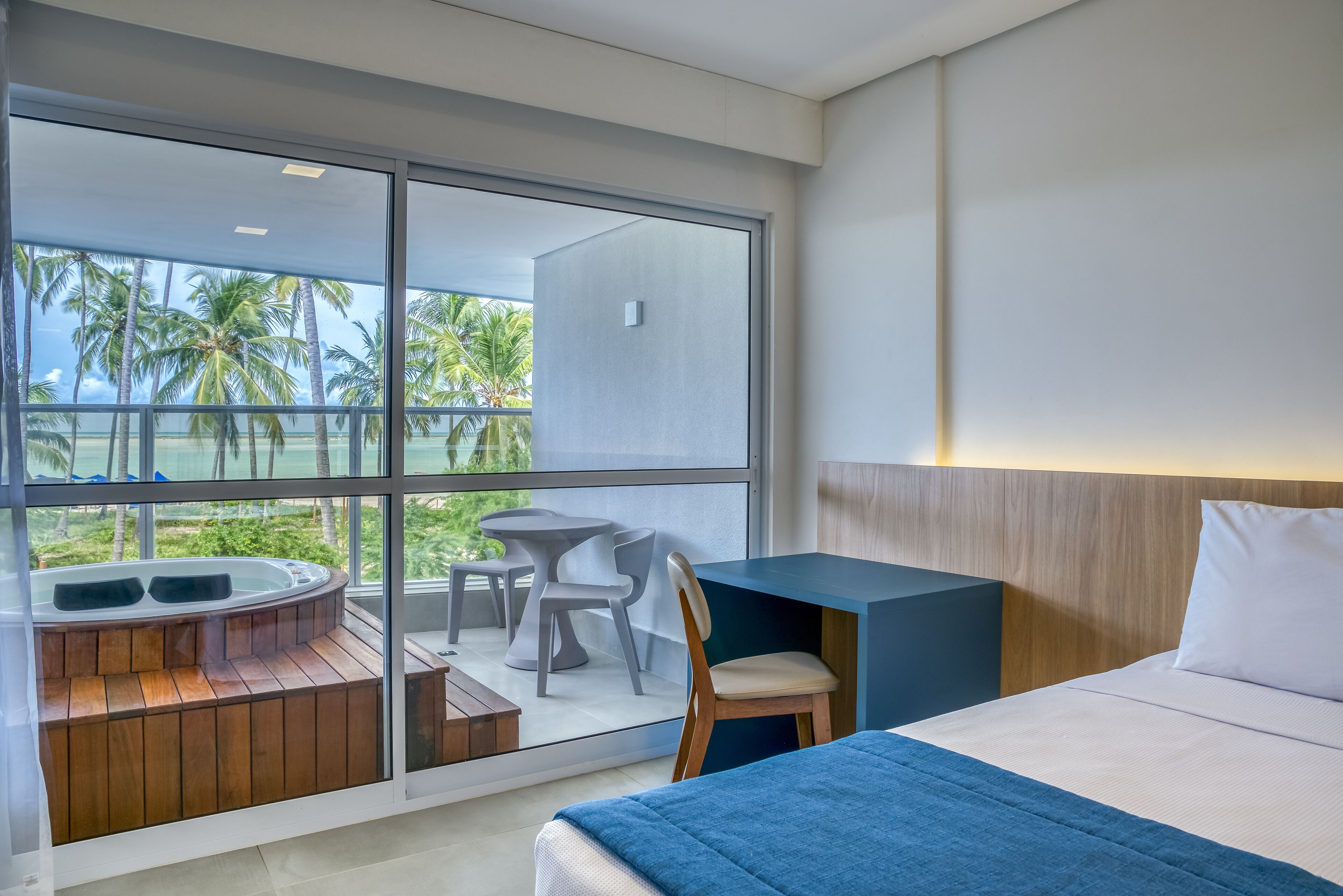 Une chambre d’hôtel moderne dotée d’une grande fenêtre, d’un bain à remous en bois, d’un lit recouvert d’une couverture bleue et d’une petite table avec des chaises.