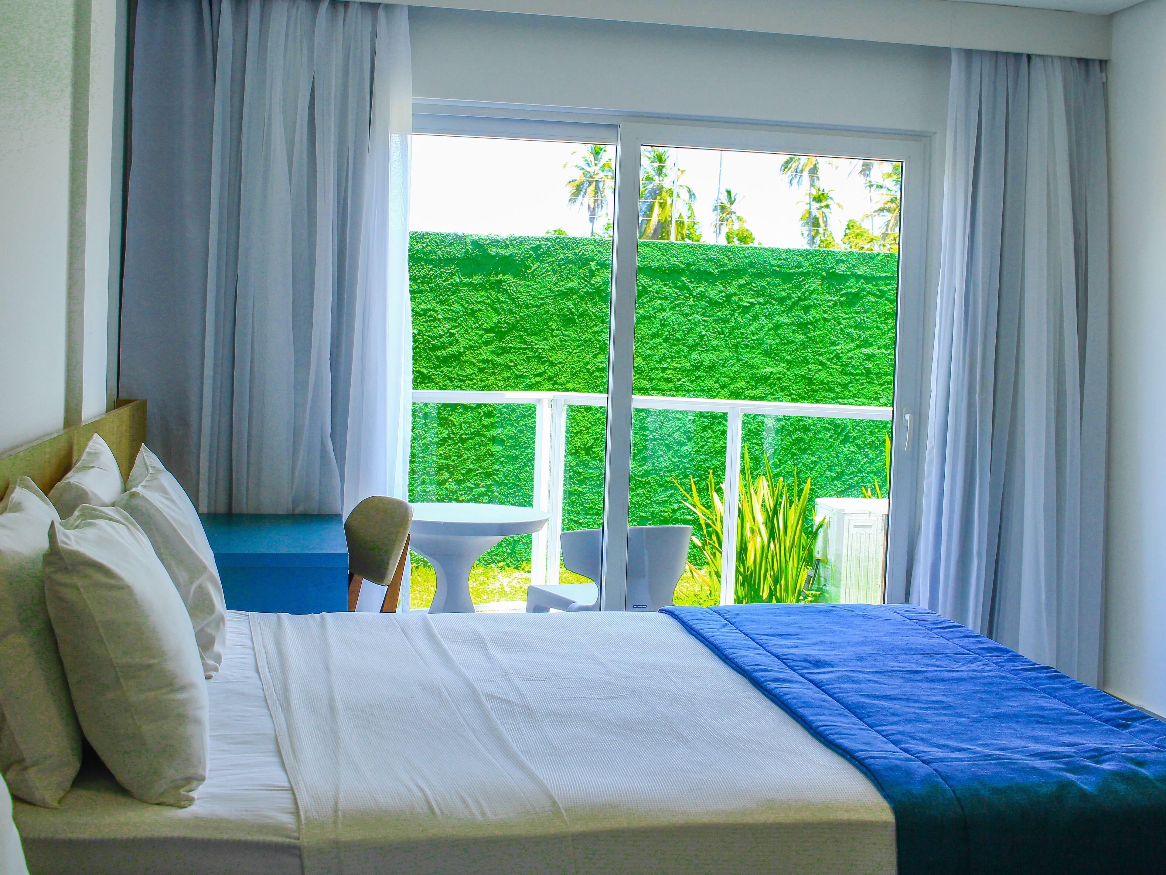 Un dormitorio con un ventanal grande, una cama con ropa de cama blanca y azul, y vistas a un seto verde al exterior.