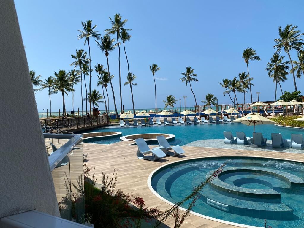 Uma área de piscina em resort com espreguiçadeiras e guarda-sóis, cercada por palmeiras.