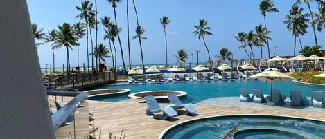 Uma área de piscina em resort com espreguiçadeiras e guarda-sóis, cercada por palmeiras.