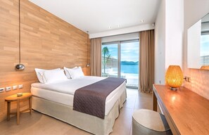 A modern hotel room with a large bed, a wooden wall, a view of the sea, and a bedside table with a lamp.