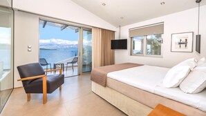 A modern hotel room with a bed, a TV, a chair, and a balcony with a view of the sea and mountains.