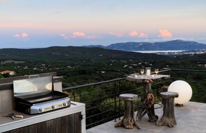 A rooftop terrace with a portable grill, a table with candles, and a view of the landscape.