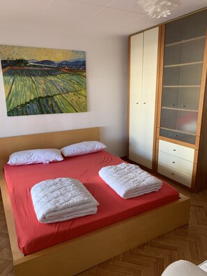 A bedroom with a bed, pillows, a red bedspread, a wooden headboard, a wardrobe with glass doors, and a painting on the wall.