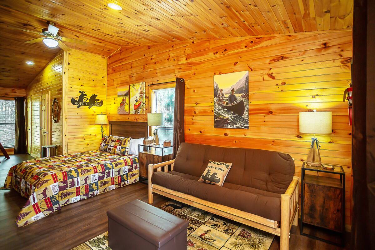A cozy cabin interior with a bed, sofa, and a small table, featuring wooden walls and a ceiling fan.