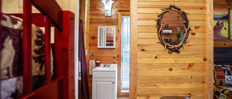 A wooden cabin interior with a bathroom, a mirror, and a bed with a colorful quilt.