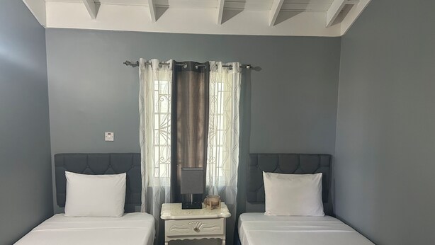 A room with two single beds, a small bedside table, a mirror, and a window with curtains. - Williams Oasis (Falmouth)