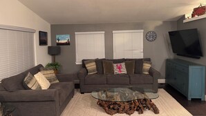 Un salón con dos sofás grises, una mesa de centro de vidrio con base de madera, un televisor en la pared y un reloj.
