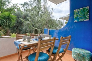 Un comedor con una mesa dispuesta para cuatro personas, rodeada de sillas de madera con cojines azules, frente a una pared azul con un cuadro de hojas tropicales.