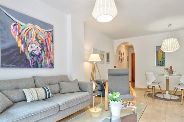 Una sala moderna con un sofá gris, una mesa de centro de vidrio y un cuadro llamativo de un león. Se observa un comedor con una mesa y sillas blancas, además de una lámpara con pantalla blanca.