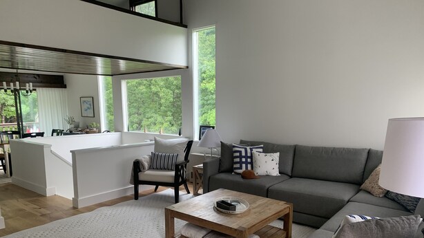 Een moderne woonkamer met een grijze bank, een houten salontafel en een fauteuil. Er is een keukenhoek met een wit aanrecht en een raam met uitzicht op bomen.