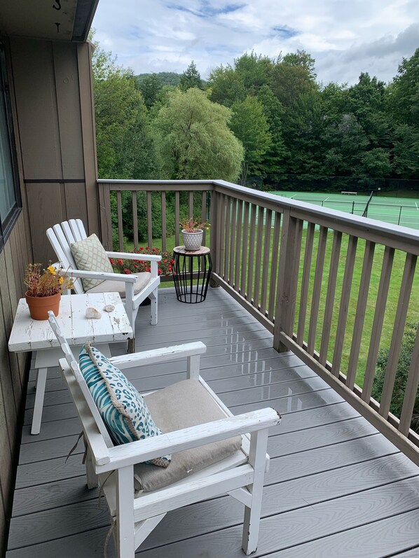 Ett trädäck med två vita Adirondack-stolar, ett litet bord med en krukväxt och utsikt över en tennisbana.