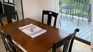 Een houten eettafel met vier stoelen, een kroonluchter, een schilderij en uitzicht op een terras met stoelen en een reling.