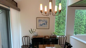 Een eetgedeelte met een houten tafel, zes stoelen, een kroonluchter en uitzicht op groen door grote ramen.