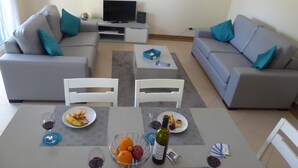 Uma sala de estar com dois sofás cinza, uma televisão e uma mesa de centro. Há duas cadeiras brancas, uma mesa de jantar com pratos de comida e copos de vinho.
