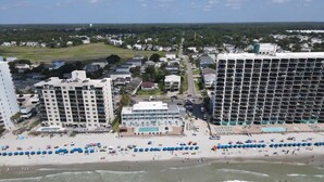 A coastal hotel with multiple floors, a beach area with umbrellas, and a view of the ocean.
