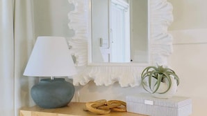 A wooden console table with a white ruffled mirror above it, a lamp, a plant, and a small stool underneath.