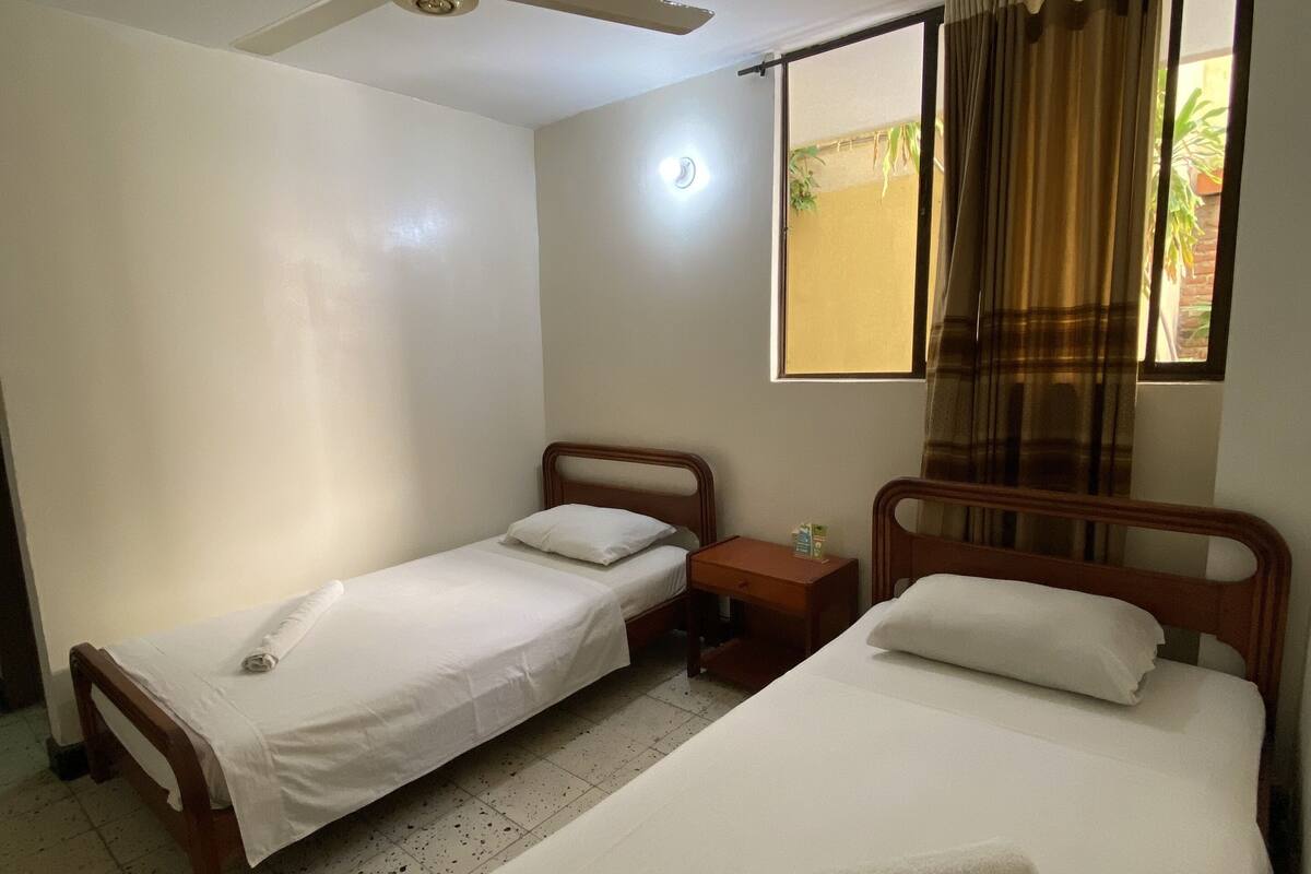 Una habitación con dos camas individuales, una mesita de noche de madera, un ventilador de techo y una ventana con cortinas.