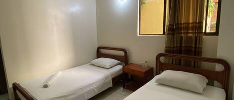 A room with two single beds, a wooden nightstand, a ceiling fan, and a window with curtains.