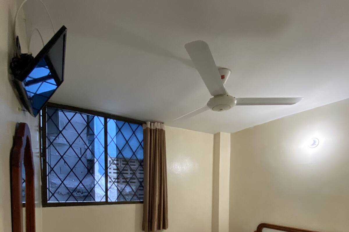 Una habitación de hotel con una cama, un ventilador de techo, una ventana con cortinas, una silla y una lámpara fijada a la pared.