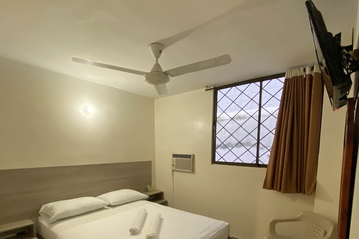 Una habitación de hotel con cama, ventilador de techo, ventana con cortinas, televisor montado en la pared y una pequeña mesa con una silla.