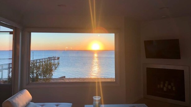 A room with a large window overlooking a sunset over the sea, a white sofa, and a small table with a cup. - Waterfront Gorgeous Beach Home-Sunsets & Spa Bath (Duck)