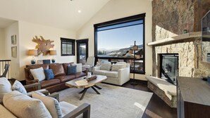 A spacious living room with a stone fireplace, large windows offering a mountain view, and a ceiling fan. - Private Hot Tub/Brand New Luxury Townhome/Short Drive to Slopes! (Keystone)