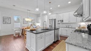 Una cocina moderna con armarios blancos, una isla central, electrodomésticos de acero inoxidable, iluminación colgante y un comedor con mesa y sillas de madera.