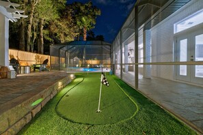 A well-lit artificial turf area with a golf hole, surrounded by a glass enclosure and a brick wall.