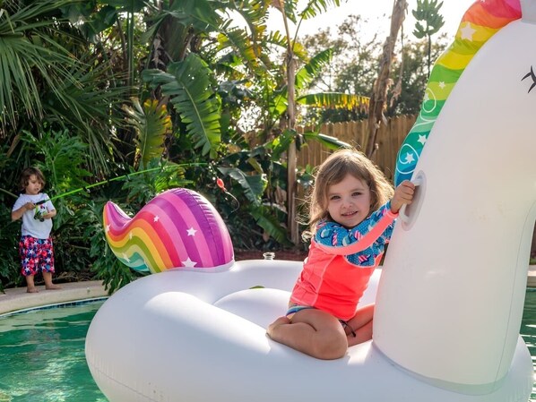 Un enfant est assis sur un flotteur gonflable en forme d’anneau, aux couleurs de l’arc-en-ciel, près d’une piscine entourée d’une végétation luxuriante.