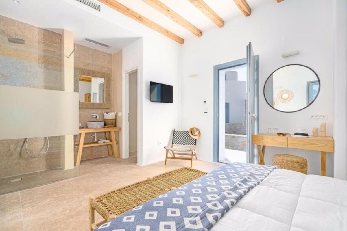 A bedroom with a bed, a wooden chair, a desk, a mirror, and a bathroom with a sink and shower.