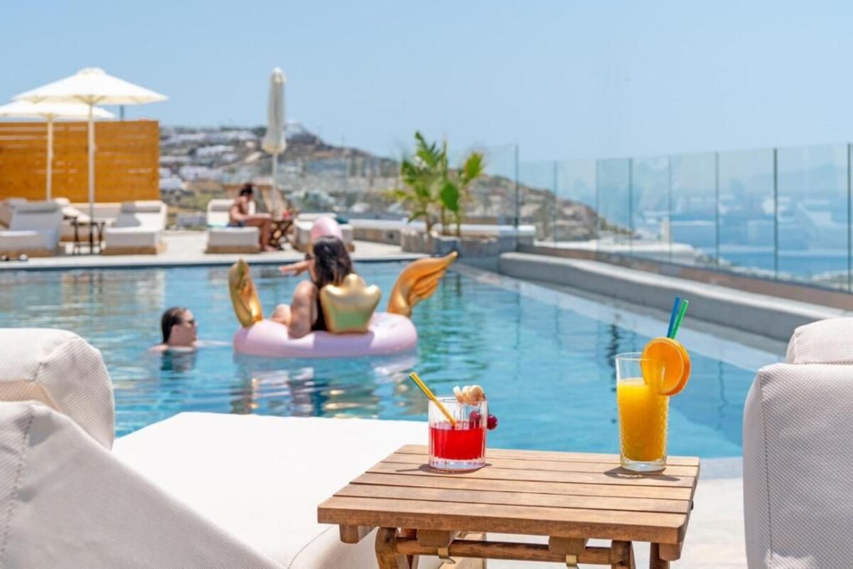 Zona de piscina con sillones de descanso, una mesa con bebidas y gente en el agua.