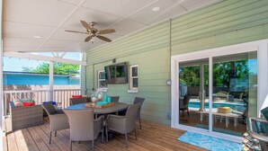 Eine überdachte Terrasse mit Deckenventilator, einem Esstisch mit Stühlen, einem an der Wand montierten Fernseher und Blick auf einen Swimmingpool durch Glastüren.