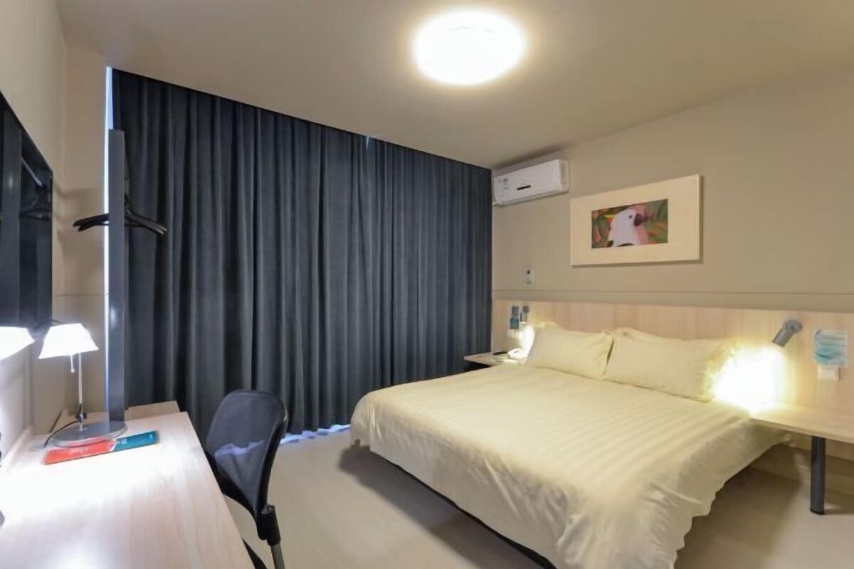 A hotel room with a bed, a desk with a lamp, a chair, a curtain, an air conditioning unit, and a framed picture on the wall.