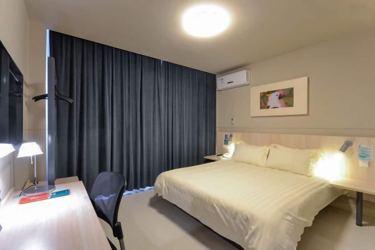 A hotel room with a bed, a desk with a lamp, a chair, a curtain, an air conditioning unit, and a framed picture on the wall.