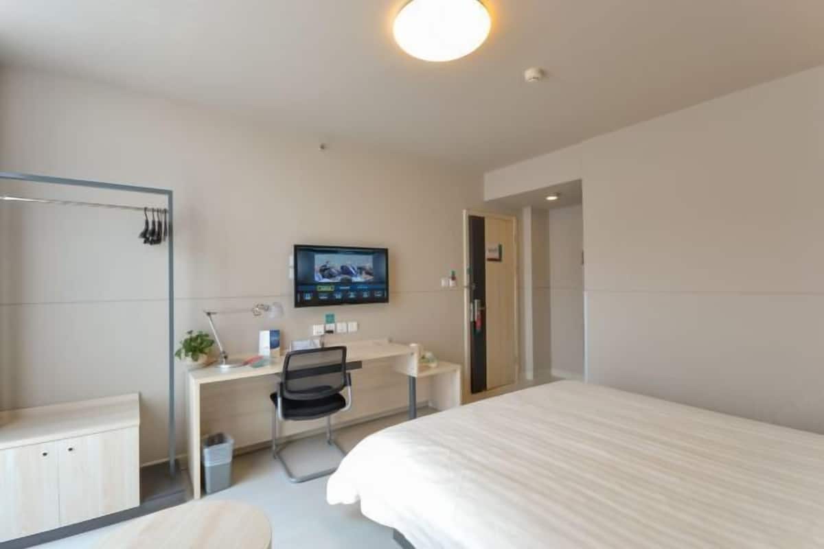A hotel room with a bed, a desk with a computer, a chair, a TV mounted on the wall, and a mirror.