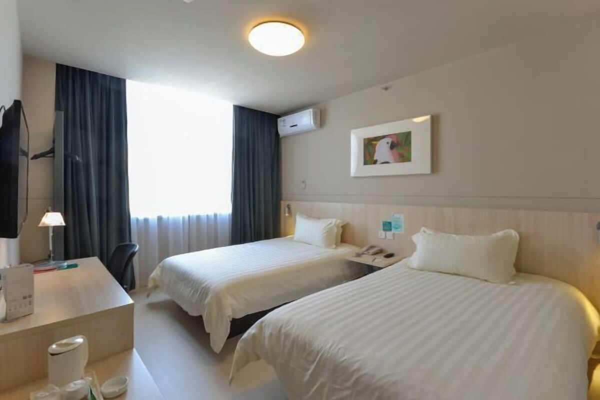 A hotel room with two beds, a desk with a lamp, a TV, and an air conditioning unit.