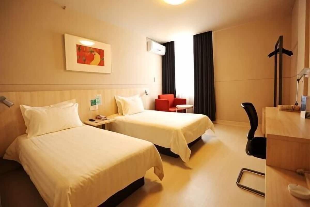 A hotel room with two beds, a desk, a chair, a small table, a painting on the wall, and an air conditioning unit.