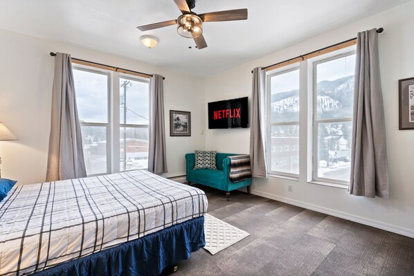A bedroom with a bed, a teal armchair, a ceiling fan, a TV mounted on the wall, and large windows with curtains.