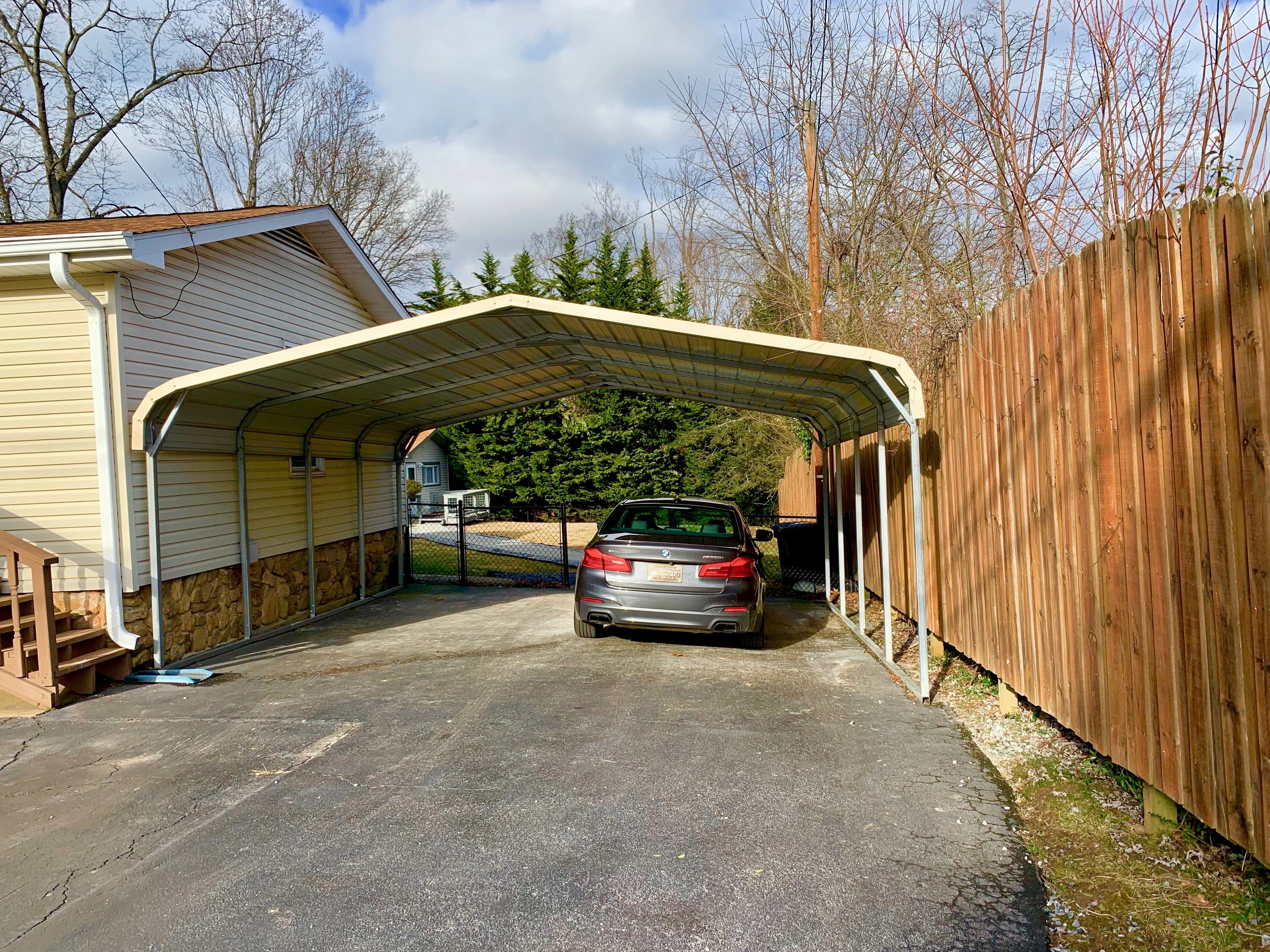 Een overdekte garage met een geparkeerde auto, een houten hek en een huis op de achtergrond.