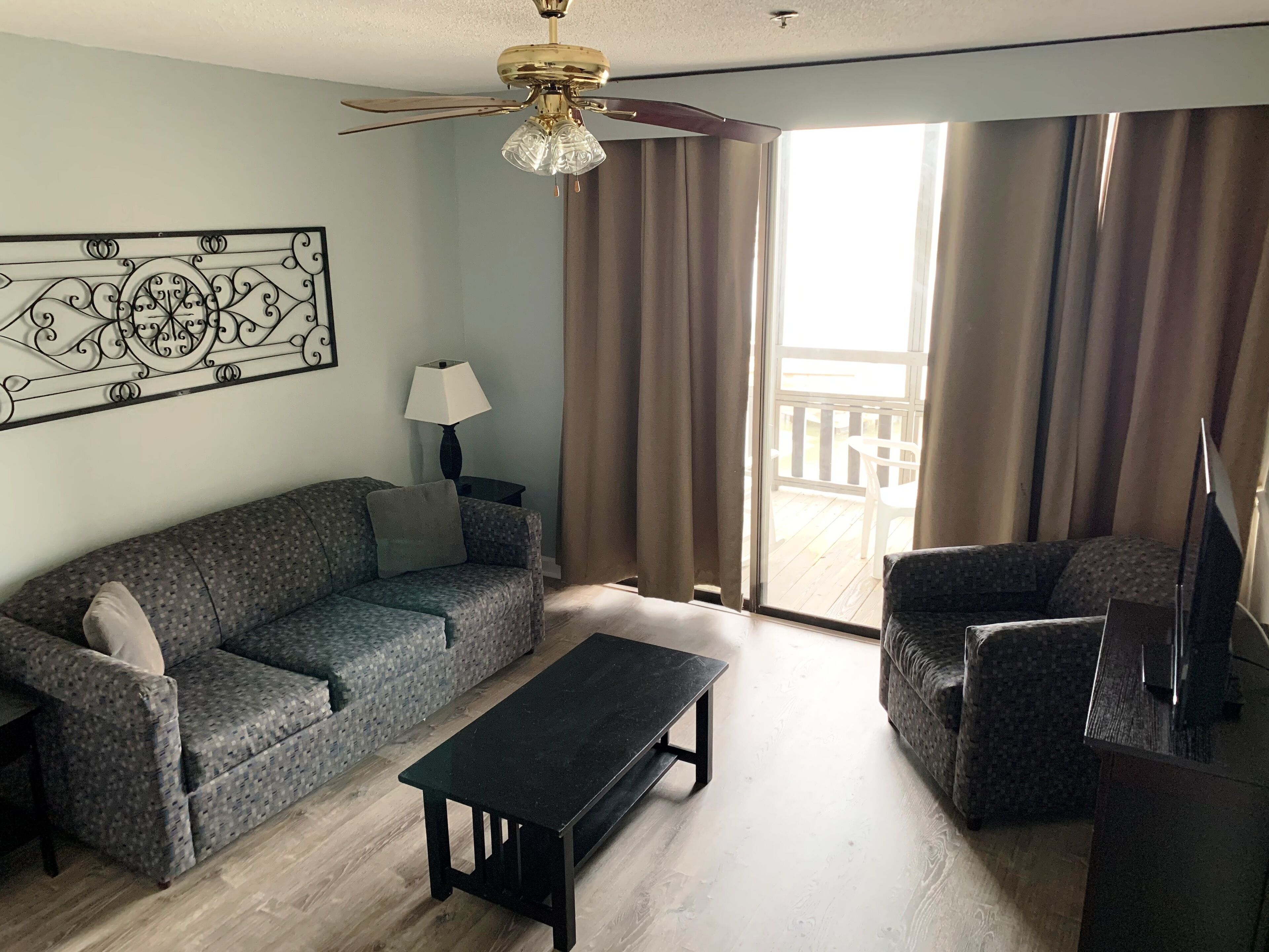 Ein Wohnzimmer mit einer Couch, einem Couchtisch, einem TV-Schrank, einem Deckenventilator und einem großen Fenster mit Vorhängen.